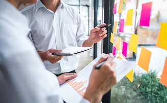 Agile Organisation Zwei Personen besprechen Diagramme und kleben bunte Post-its an eine Glaswand