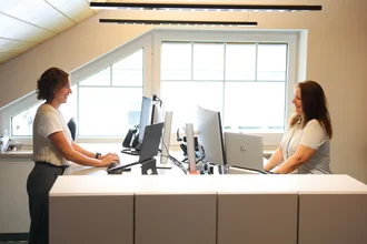 1C1A1031 Zwei Mitarbeitende im Büro stehen an einem höhenverstellbaren Doppel-Schreibtisch und arbeiten am Computer; heller Raum mit großen Dachfenstern.
