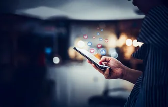 Social Media Ads Person tippt auf ein Smartphone, über dem Social-Media-Icons wie Herzchen und Likes schweben – Symbolbild für Interaktion in sozialen Netzwerken