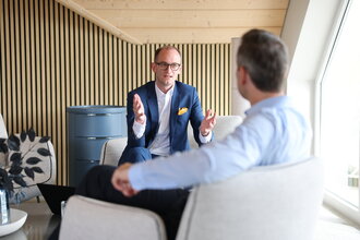 Consulting Beratungsgespräch bei Denkströme: Ein Manager im Anzug erklärt gestikulierend etwas in einem Lounge-Bereich, gegenüber sitzt ein Gesprächspartner.