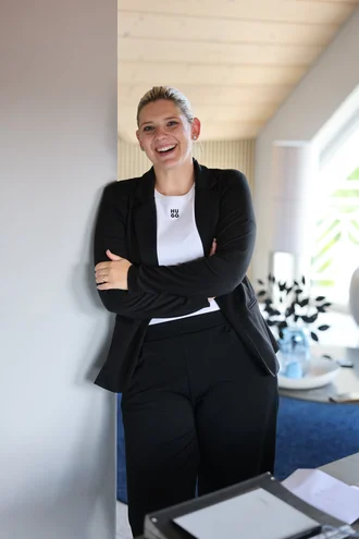Teresa Schmid von Denkströme steht lächelnd im Büroflur, lehnt an einer Wand und hat die Arme verschränkt.