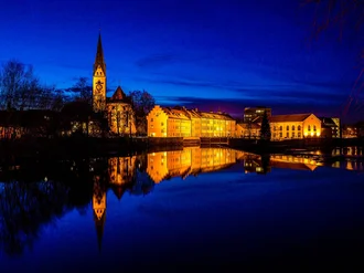 Abendliche Stadtansicht von Kempten mit beleuchteten historischen Gebäuden und Kirche am Wasser, gespiegelt im ruhigen Fluss – symbolisiert Standortverbundenheit, Tradition und Atmosphäre im Allgäu