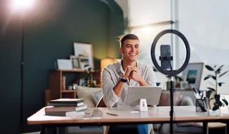 Social Media Content Junger Mann sitzt an einem Schreibtisch mit Tablet, lächelt in eine Kamera, die auf einem Stativ mit Ringlicht montiert ist – moderne Content-Produktion oder Videokonferenz in gemütlichem Büro