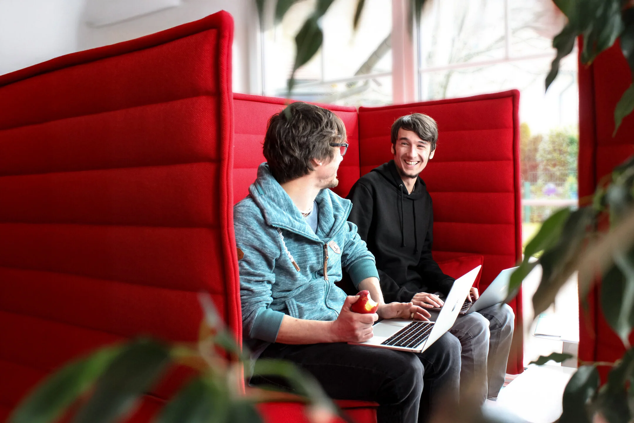 Zwei Denkströme Mitarbeiter im Gespräch in modernem Büro mit roten Akustik-Sesseln, arbeiten gemeinsam mit Laptops, kreativer Austausch