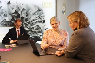 Projektmanagement / -leitung Denkströme Geschäftsführerin und Mitarbeiterin im Gespräch vor Laptop in modernem Büro – Teammeeting mit Fokus auf digitale Zusammenarbeit, im Hintergrund Denkströme Geschäftsführer mit Tablet