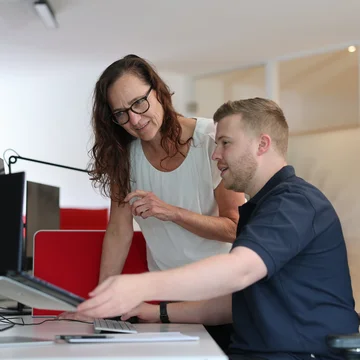 Zwei Mitarbeitende aus der Redaktion von Denkströme arbeiten am Schreibtisch: Eine Person zeigt auf den Monitor, die andere sitzt am Computer und hört zu.