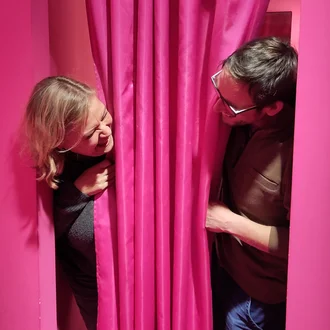 Zwei Teammitglieder von der Denkströme blicken sich lachend durch einen pinken Vorhang an – eine Szene voller Humor und Teamgeist