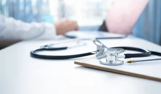 Prozessdigitalisierung Stethoskop auf einem Schreibtisch mit Klemmbrett und Kugelschreiber, im Hintergrund eine Person am Laptop – Szene aus einem medizinischen Arbeitsumfeld
