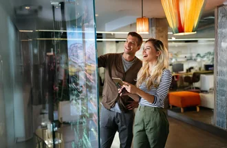 Social Media Recruiting Zwei lächelnde Menschen – ein Mann und eine Frau – stehen in einem modernen Büro vor einem großen Glas-Whiteboard, auf dem mit bunten Markern Brainstorming-Notizen geschrieben sind. Die Frau hält ein Tablet und eine Brille in der Hand.